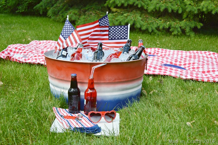 Red, white, and blue striped party tub filled with ice and bottles near a picnic blanket.