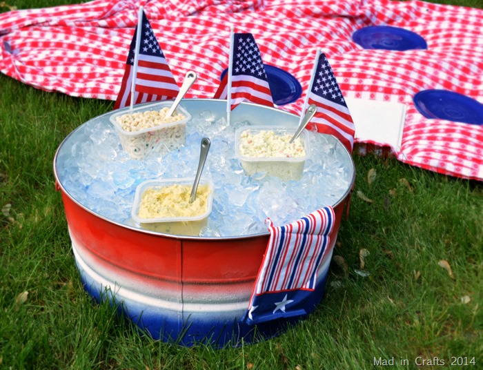 Red, white, and blue striped party tub filled with ice and picnic salads near a picnic blanket.