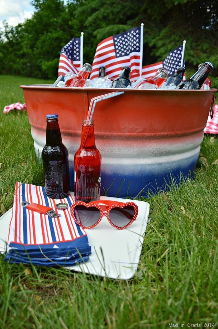 Closeup of a red, white, and blue striped party tub filled with ice and bottles near a picnic blanket.