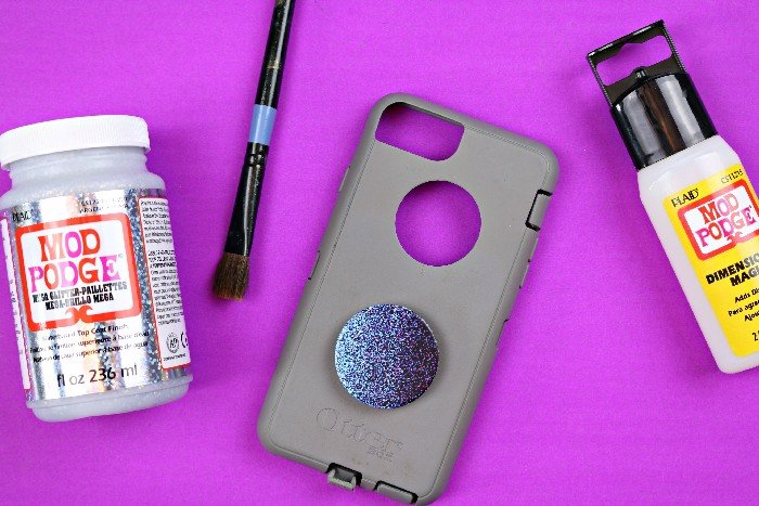 Flay lay of Mega Glitter Mod Podge, a paintbrush, a phone case with a Pop Socket, and Mod Podge Dimensional Magic