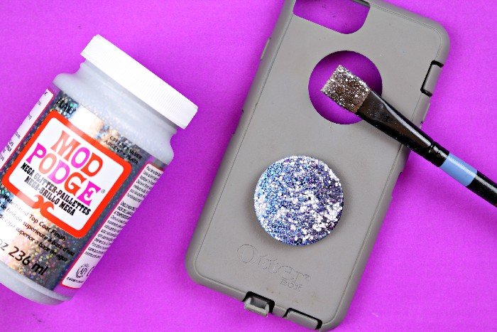 A bottle of Mega Glitter Mod Podge next to a phone case with a Pop Socket on it and a paintbrush.