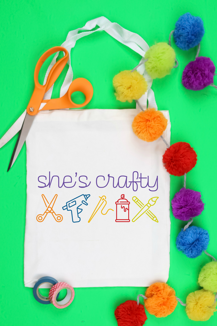 she\'s crafty SVG tote bag, fabric scissors and rainbow pom garland on a green background