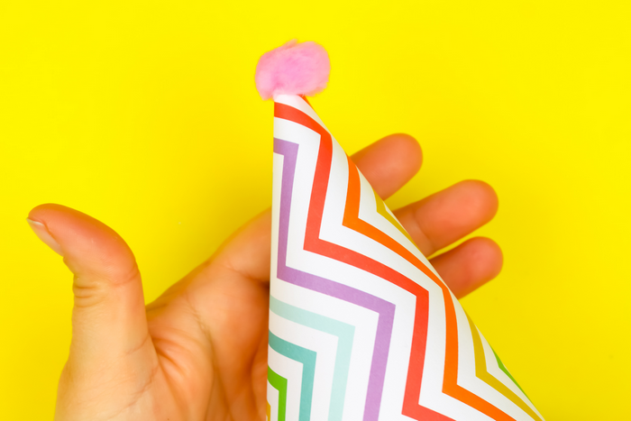 hand holding a colorful paper party hat in front of a yellow background