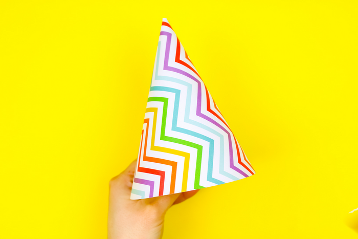Hand holding a colorful paper party hat in front of a yellow background