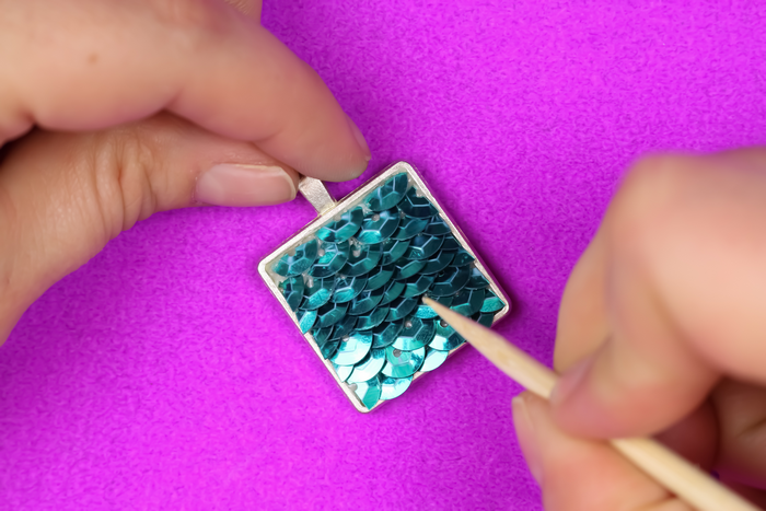hand holding a jewelry bezel with teal sequins and a toothpick on a purple background