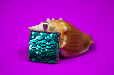 teal sequin mermaid pendant leaning on a seashell on a purple background