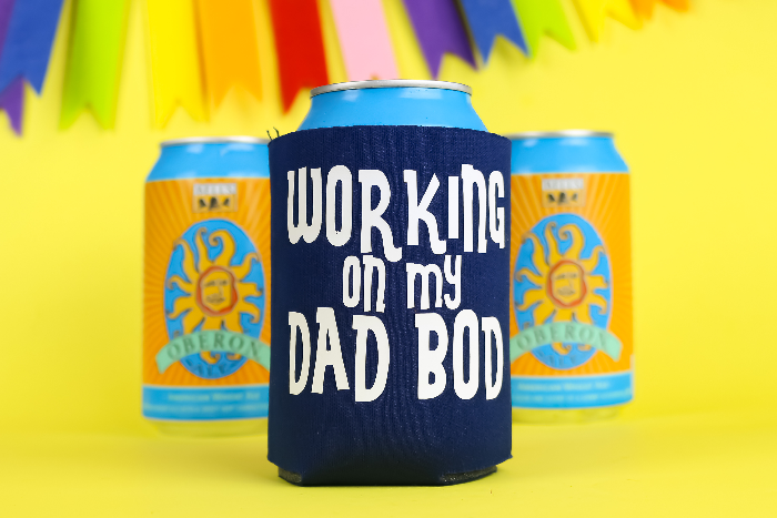 Navy can kooziethat says Working on My Dad Bod in white vinyl next to 2 cans of beer