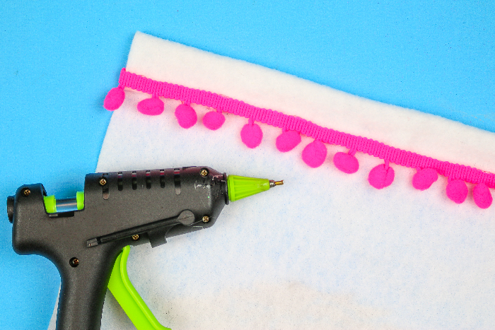 pink pom pom trim on white felt on a blue background with a glue gun
