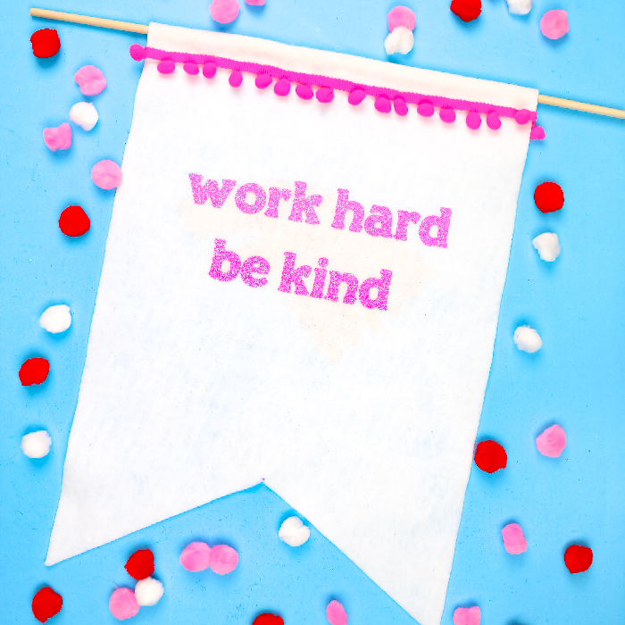 white felt banner that says work hard be kind on a blue background with pom poms