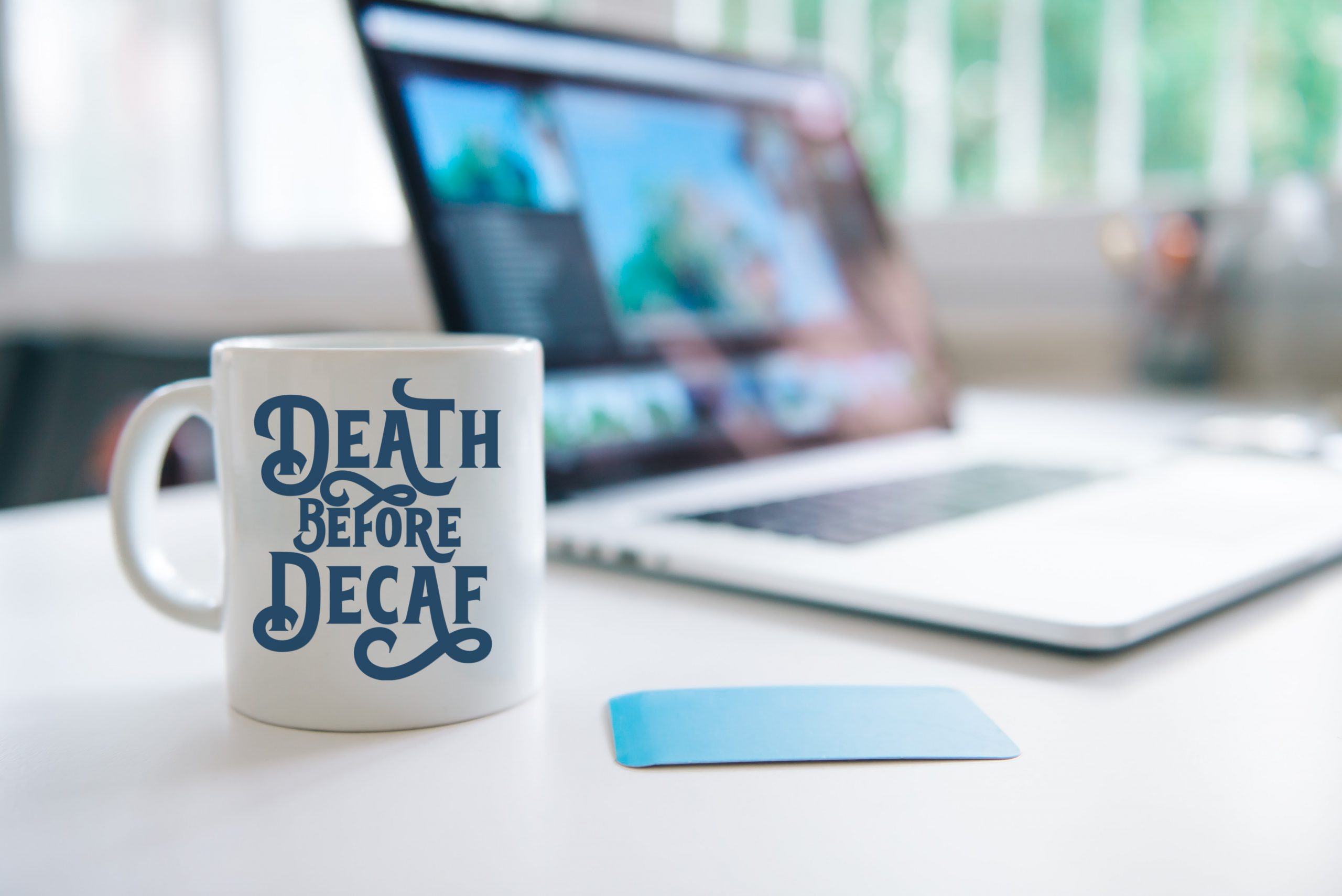 white mug that reads Death Before Decaf on a desk next to a laptop