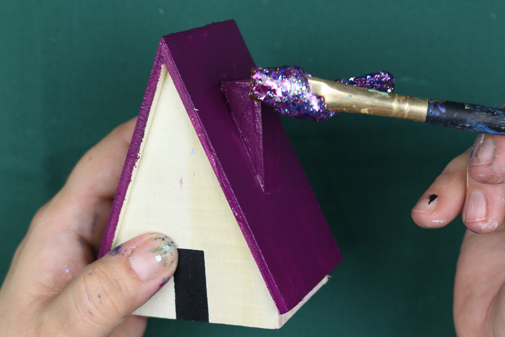 hands applying purple glitter paint to the roof of a wooden house