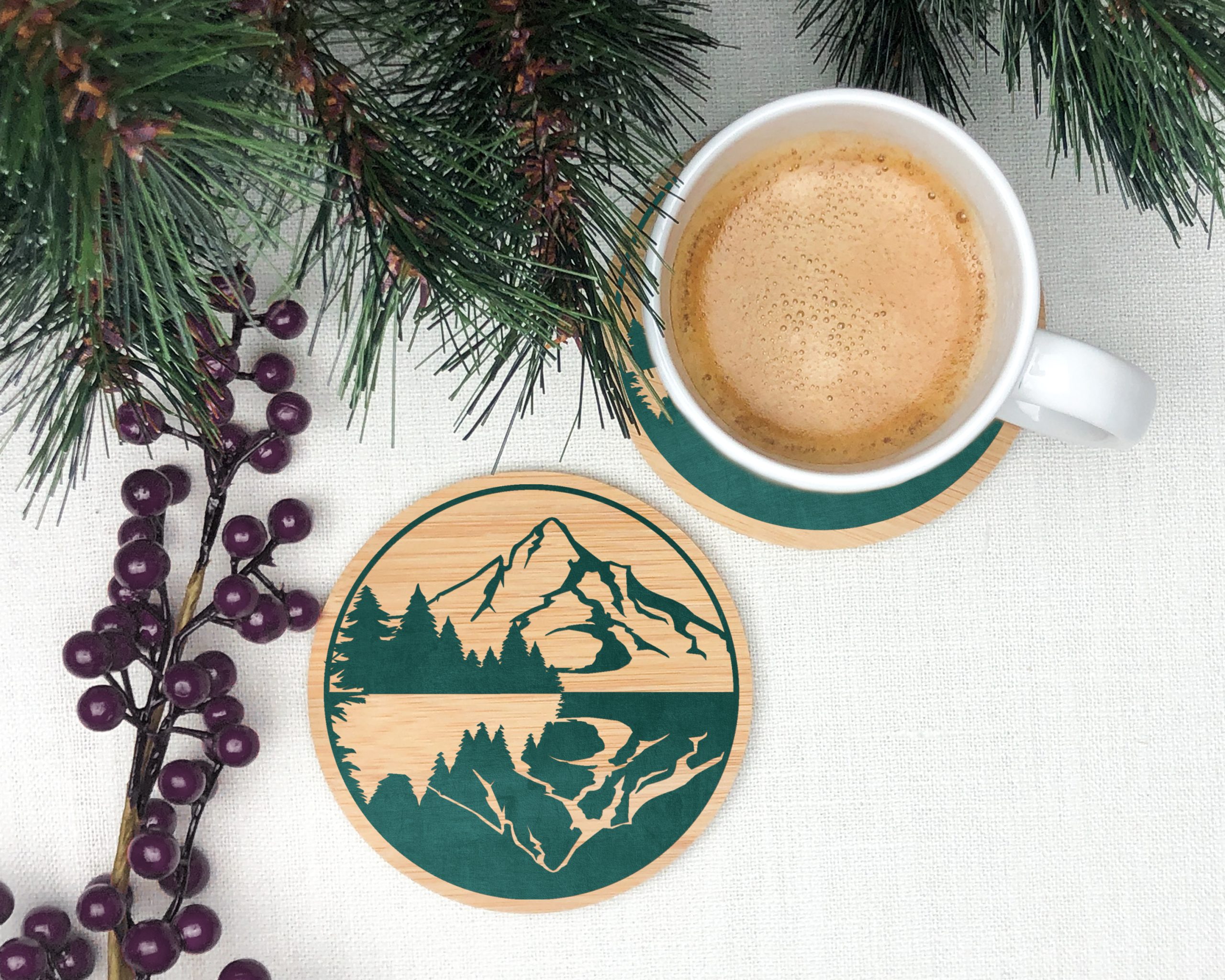 wood coaster decorated with a vinyl mountain scene near a mug o coffee and pine branches
