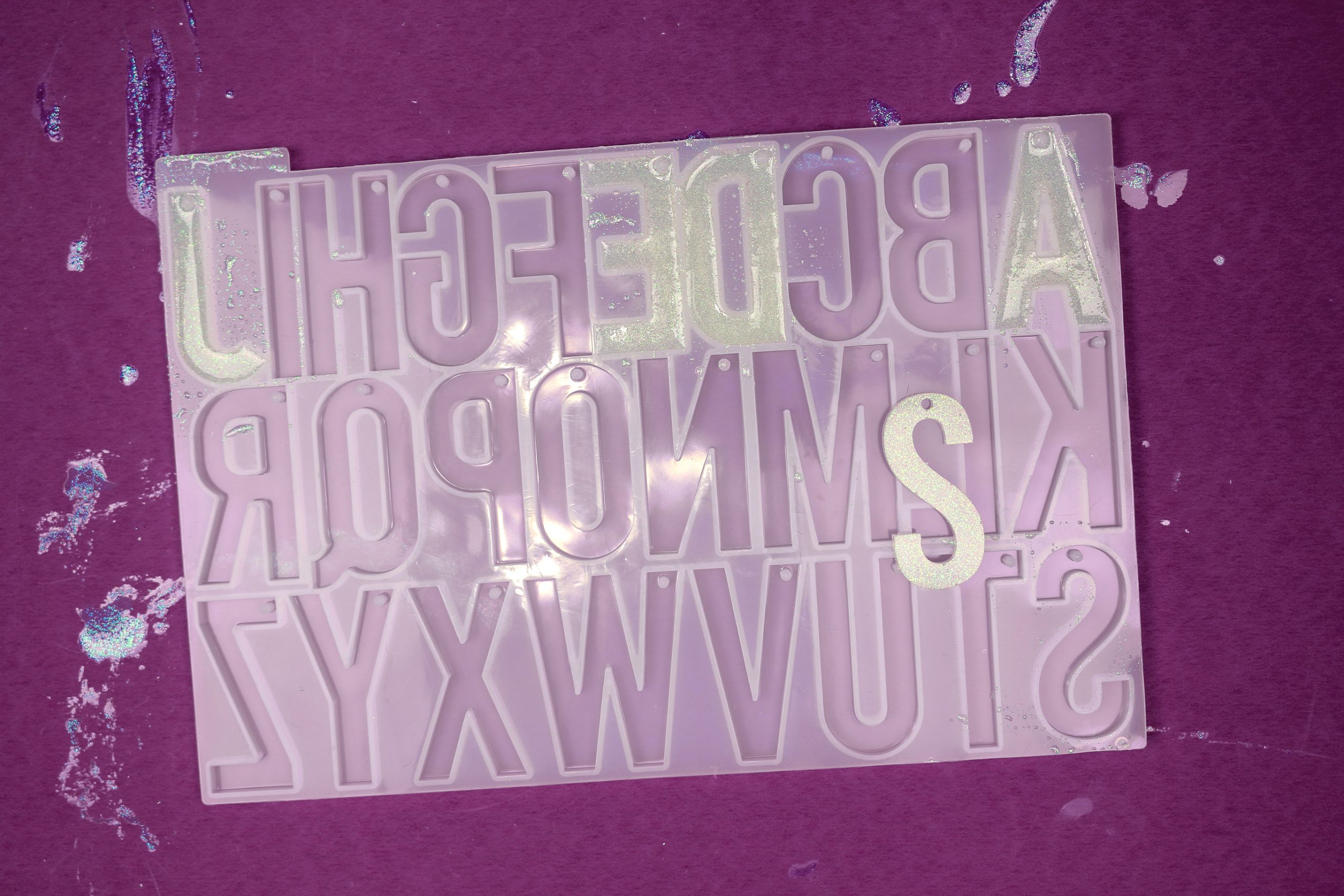 Silicone alphabet mold with a few cavities filled with glittery white resin. One letter has been removed from the mold
