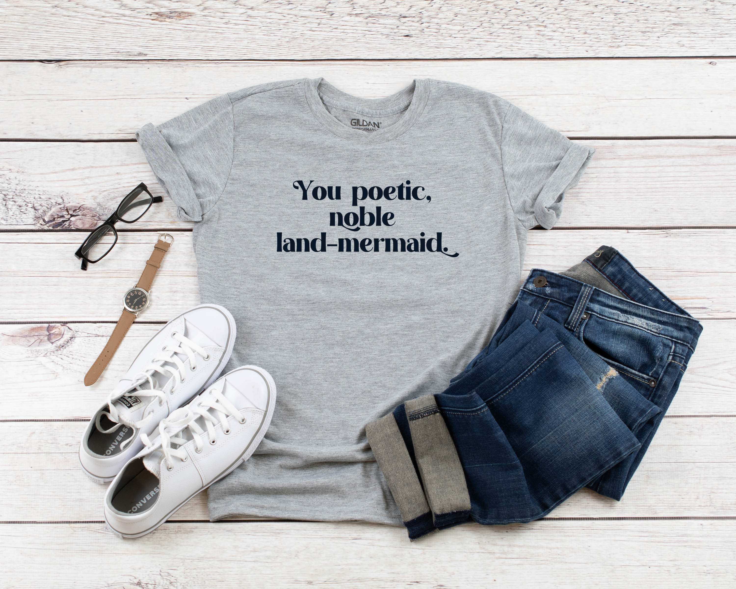 grey t-shirt that reads You poetic, noble land mermaid sits near white shoes and blue jeans