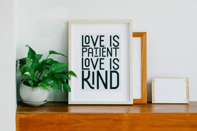Woo frame containing a print that reads "Love is Patient Love is Kind" on a shelf near a plant