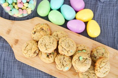 Top view of a pile of Cadbury Mini Egg Cookies on a wood cutting board near plastic eggs and a bowl of Cadbury Mini Eggs.