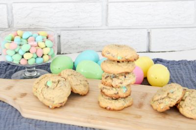 Side view of a pile of Cadbury Mini Egg Cookies on a wood cutting board near plastic eggs and a bowl of Cadbury Mini Eggs.