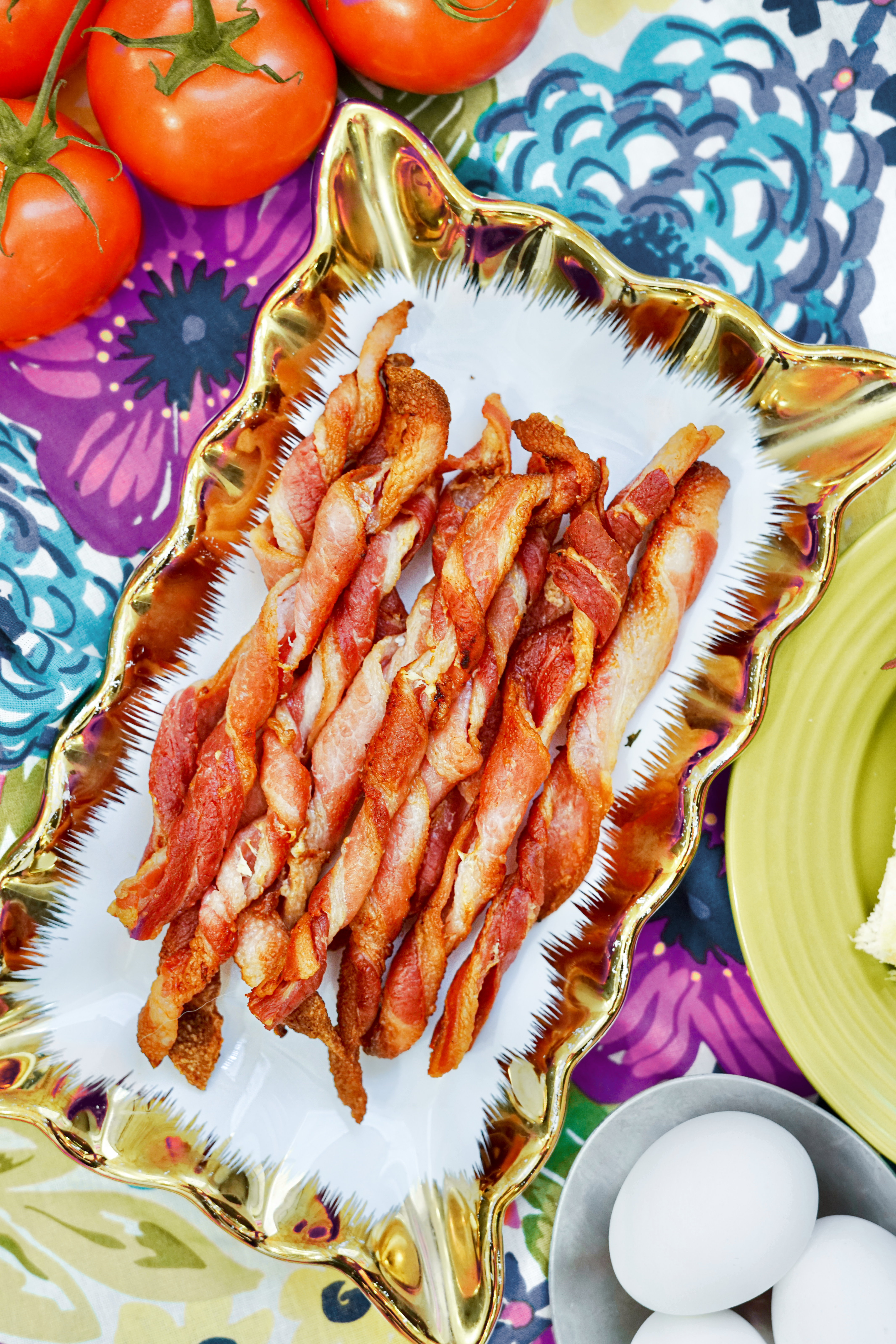 Platter of air fried bacon twists on a colorful tablecloth near tomatoes and eggs.