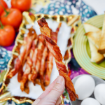 Woman's hand holding one bacon twist in front of Platter of air fried bacon twists on a colorful tablecloth.