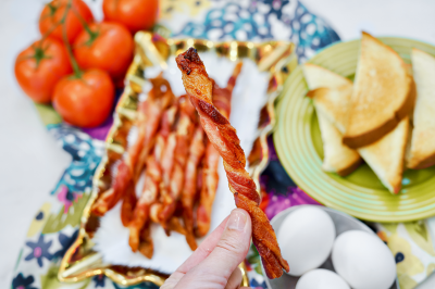 Woman's hand holding one bacon twist in front of Platter of air fried bacon twists on a colorful tablecloth.