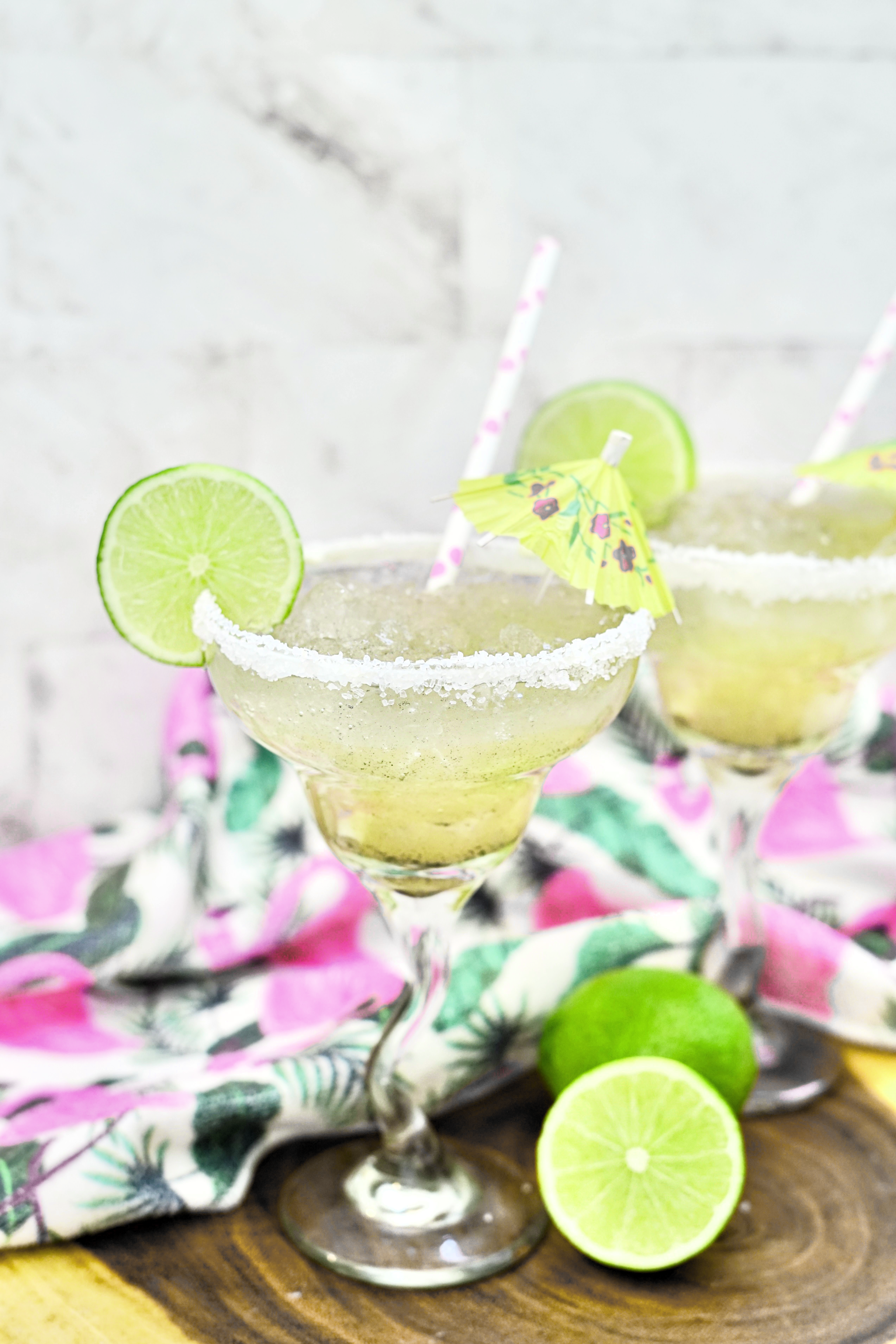 A frozen lime margarita with salt rims that has been garnished with lime, a paper straw, and a small umbrella.