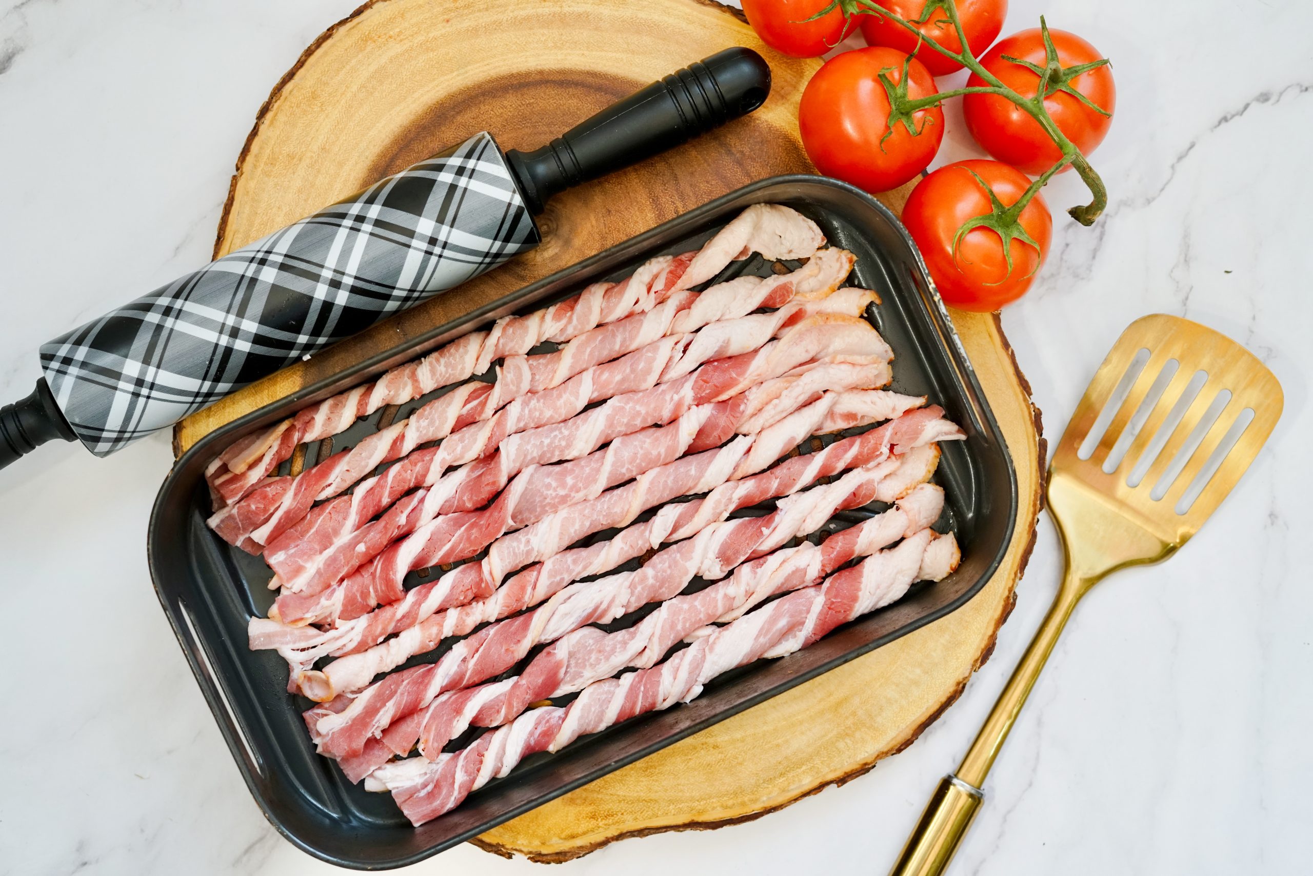 Uncooked bacon twists in an air fryer tray near kitchen tools.