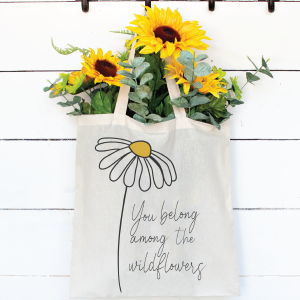 Canvas tote bag holding yellow flowers. The tote bag is decorated with a You Belong with the Wildflowers SVG