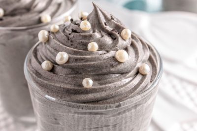 A closeup of a glass jar is filled with piped and swirled grey stuff dessert topped with white sprinkles
