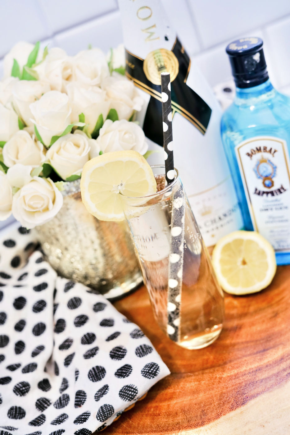 An angled shot of a French 75 cocktail near a vase of white roses and bottles of champagne and gin.