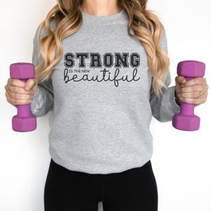 A white woman holds dumbells and wears a great sweatshirt that reads Strong is the new beautiful