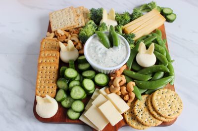 snack board covered with green vegetables, cheese, crackers and a bowl of ranch dip