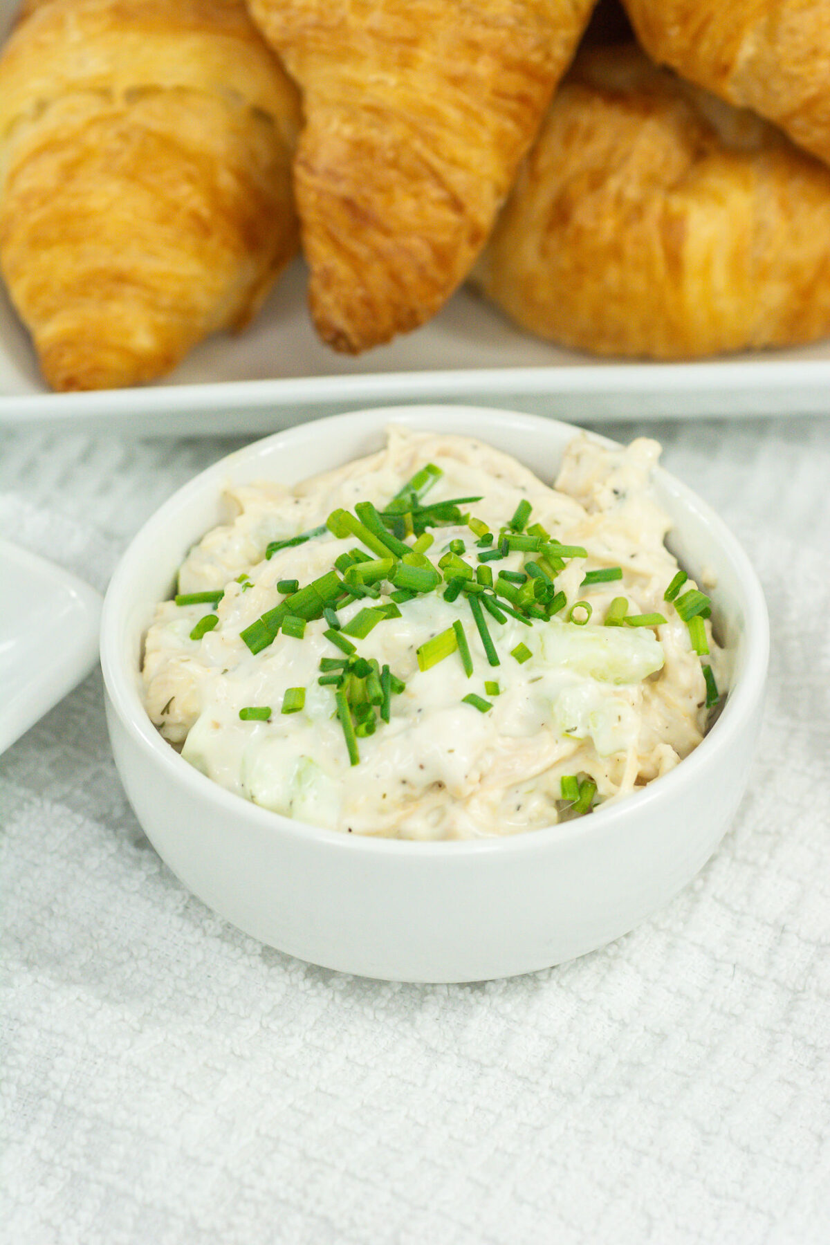Closeup of a white bowl of chicken salad near a platter of croissants