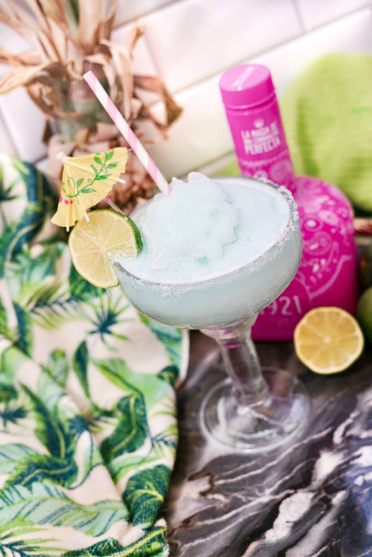 tilted view of frozen blue margarita garnished with a lime slice near a bottle of tequila