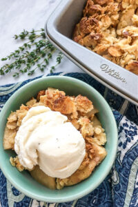 A green bowl full of apple crisp topped with vanilla ice cream near a pan of crisp.