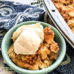 A closeup of a green bowl full of apple crisp topped with vanilla ice cream near a pan of crisp.