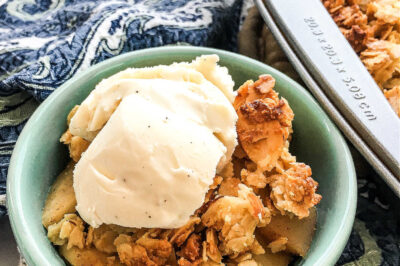 A closeup of a green bowl full of apple crisp topped with vanilla ice cream near a pan of crisp.