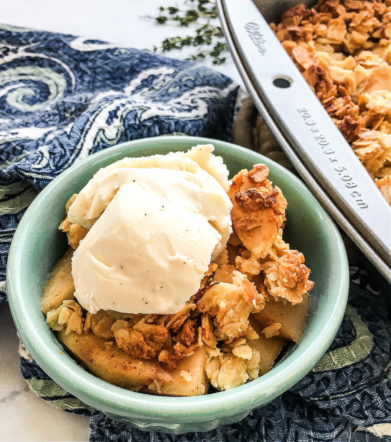 A closeup of a green bowl full of apple crisp topped with vanilla ice cream near a pan of crisp.