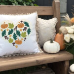 fall heart design on a white pillow sitting on an outdoor bench near pumpkins and mums