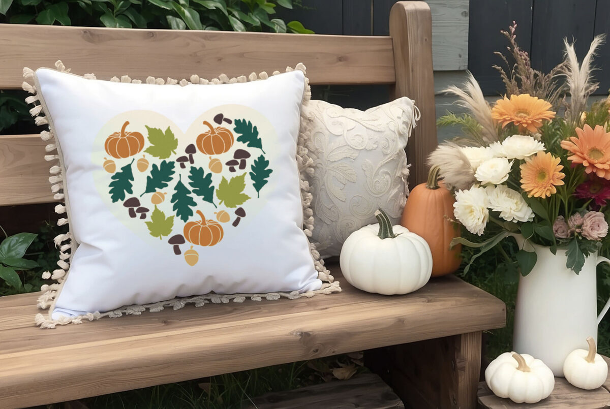 fall heart design on a white pillow sitting on an outdoor bench near pumpkins and mums