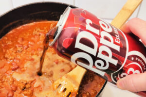 white woman's hand pouring Dr. Pepper into a pot of chili